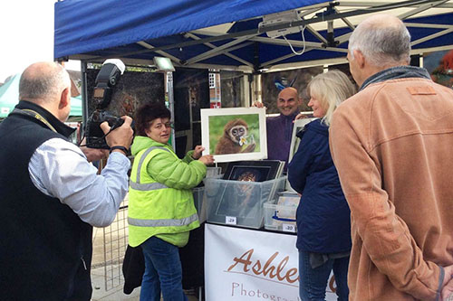 lady in a green high-visibility jacket holding up one of Ashley Vincent’s wildlife prints in from of his event stand while a news reporter takes their photo