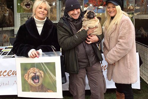 lady customer in front of event stand holding a picture of a lion cub she has just purchased from the photographer Ashley Vincent, with her friend looking on and Ashley holding her dog